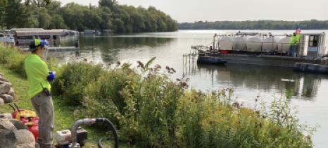 Pumps and hoses are on the lakeshore where a man watches a barge with white plastic tanks being filled