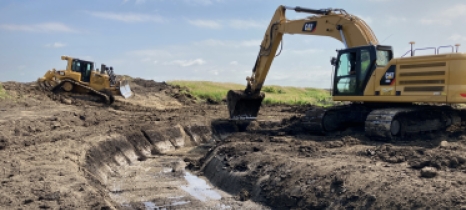 A backhoe excavates a stream channel. A bulldozer is in the background.