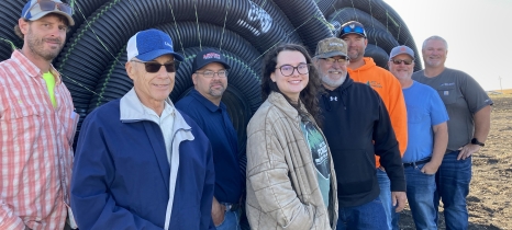 eight people stand in front of rolls of black plastic tile with a bare field in the background