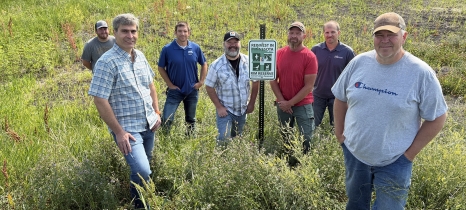 Seven people stand in tall plants around a Reinvest in Minnesota Reserve sign.